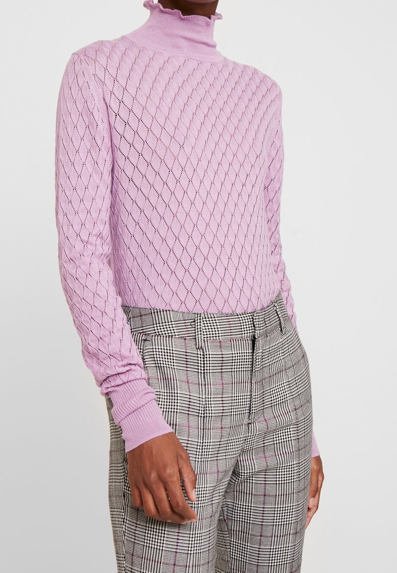 Un pulls en tricot rose clair avec un motif diamant texturé, doté d'un col haut à volants et de manches longues. Associé à un pantalon à carreaux.