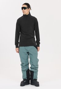 Schwarzes Langarmoberteil mit einem Reißverschlusskragen, kombiniert mit helltürkisfarbenen Schneehosen. Schwarze Stiefel vervollständigen das Winter-Sportoutfit.
