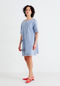 Denim obleka, svetlo modra, kratki rokavi, širok kroj, stranske vezave, dolžina nad kolenom. Model nosi rdeče balerinke. Enostaven in funkcionalen dizajn.