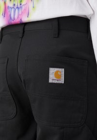 Zwarte cargobroek van stevig stof, met meerdere zakken en stiksels. Beschikt over een wit label met een geel logo.