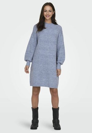 Vestido de punto azul con un cuello alto, mangas largas abullonadas y un patrón texturizado. Llevado con botines negros hasta el tobillo. Longitud hasta el muslo.