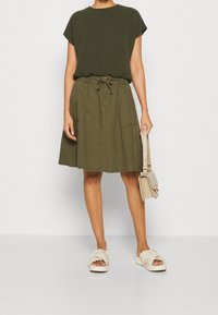 Top de manga corta texturizado en verde oliva con cuello redondo, combinado con una falda a juego con cintura elástica y bolsillos laterales. Bolso en tonos neutros y sandalias claras.