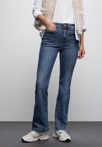 Jeans a gamba larga blu in denim con due tasche frontali, cuciture sottili e vita a media altezza, abbinati a scarpe da ginnastica di colore chiaro.