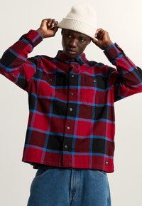 Rood, zwart en blauw geruite flanellen shirt met twee voorzakken en een knoopsluiting bij de kraag. Model draagt een crème beanie. Spijkerbroek zichtbaar.