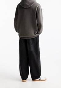 En person som bär en mörkgrå hupptröja, lösa svarta jeans och vita sneakers står med händerna i fickorna, vänd bort.