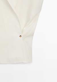 Tissu ivoire avec un petit bouton rond en or visible sur un vêtement plié, présenté sur un fond blanc.