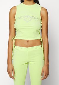 Femme portant un crop top sans manches vert citron avec un motif cœur en strass et un pantalon assorti avec des détails lacés sur les côtés.