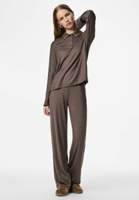 Žena u smešnoj smešnoj duljini s dugim rukavima i odgovarajućim širokim hlačama, stoji s jednom rukom iza vrata nasuprot bijeloj pozadini.