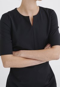 Svart klänning av mjukt tyg med kort ärm och en subtil nyckelhålsdetalj vid halsen. Modellens armar är korsade.