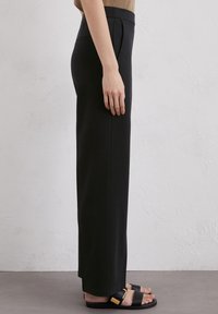 Schwarze, maßgeschneiderte Hose mit geradem Bein, hoher Taille und Seitentaschen, kombiniert mit schwarzen Sandalen und einem strukturierten Oberteil.