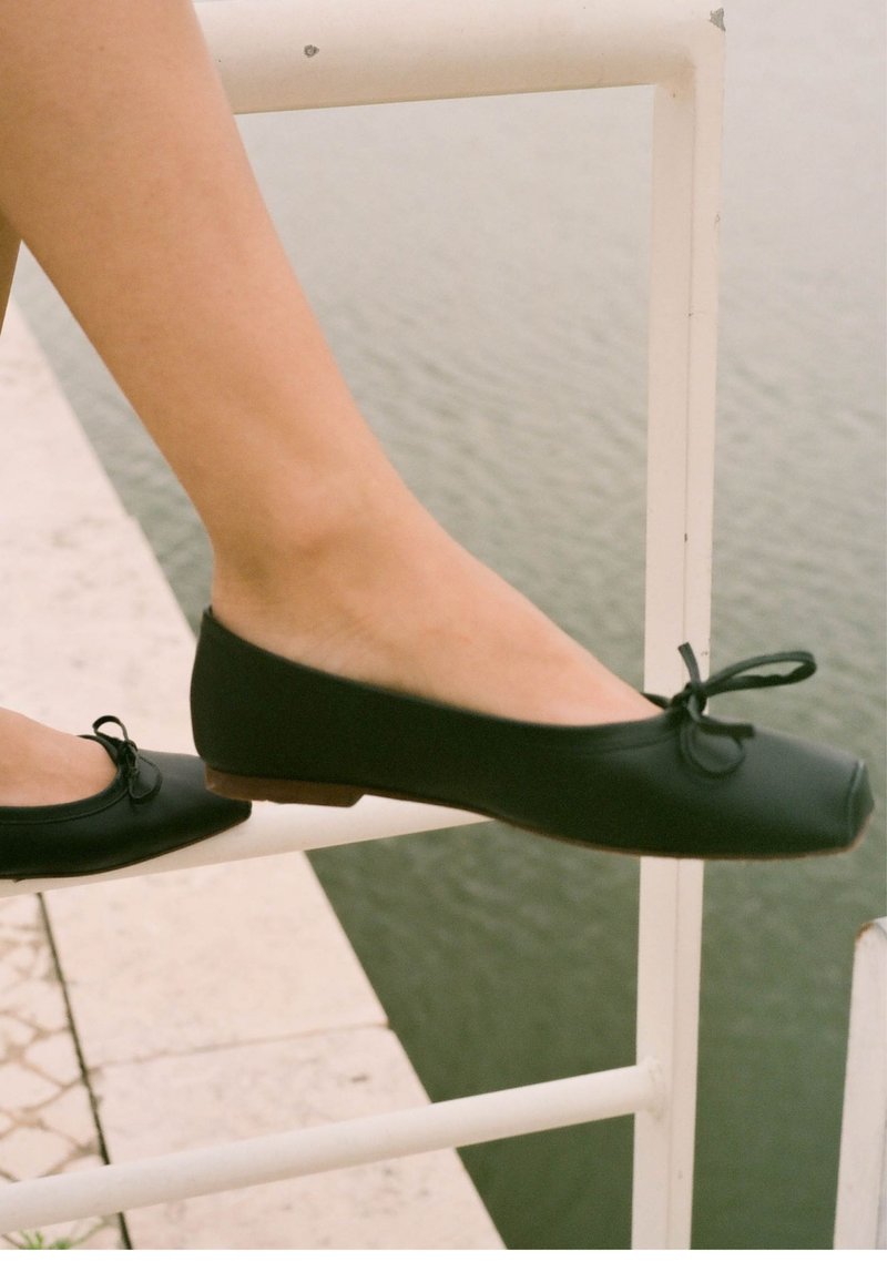 Zapatos planos negros con punta redondeada, fabricados en material suave, con un pequeño detalle de moño en la parte frontal y un bajo tacón de madera marrón.