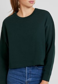 Ciemnozielona krótka bluza z długimi rękawami i okrągłym dekoltem, wykonana z miękkiego materiału. Charakteryzuje się luźnym krojem i prostym wzorem.