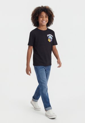 Smilende barn med krøllet hår, iført en sort Pokémon t-shirt, blå jeans og hvide sneakers mod en ensfarvet lys baggrund.