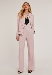 Blazer su misura rosa chiaro con bottoni neri, abbinato a pantaloni a gamba larga dello stesso colore. Sotto c'è una maglietta nera con dettagli in pizzo.
