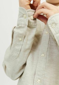 Camicia a maniche lunghe di un beige chiaro con bottoni visibili e dettagli sui polsini cuciti. Il tessuto appare morbido e testurizzato. I bottoni sono rotondi e bianchi.