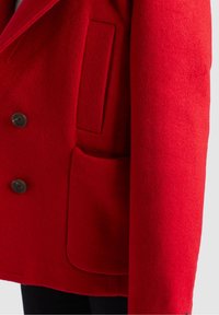 Manteau en laine rouge avec des boutons noirs, doté d'une poche à rabat et d'une poche passepoilée verticale sur le côté gauche.