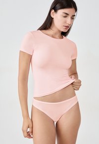 Frau in passendem, hellrosa kurzärmeligen Oberteil und Unterwäsche, die sanft den Saum des Oberteils anhebt, vor einem schlichten Hintergrund.