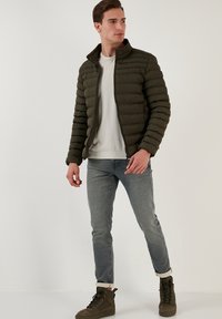 Oliven grøn polstret jakke med opretstående krave, lynlås og vandret syninger. Kombineret med lyseblå jeans og mørke sneakers.