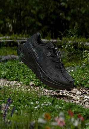 Scarpa da trail running Salomon nera con suola testurizzata, che fluttua sopra un sentiero roccioso circondato da fogliame verde e piccoli fiori.