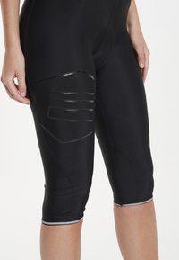 Schwarze sportliche Capri-Hosen mit glatter Textur, die über glänzende Einsatzdetails an den Oberschenkeln und reflektierende Akzente am Saum verfügen.