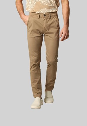 Chinos beiges avec une coupe slim, fermeture à bouton et texture subtile. Associés à des chaussures slip-on de couleur claire et une chemise à motifs.