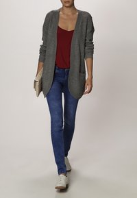 Woman wearing gray knit cardigan, red top, blue jeans, white shoes, and holding beige clutch, walking forward on plain background.