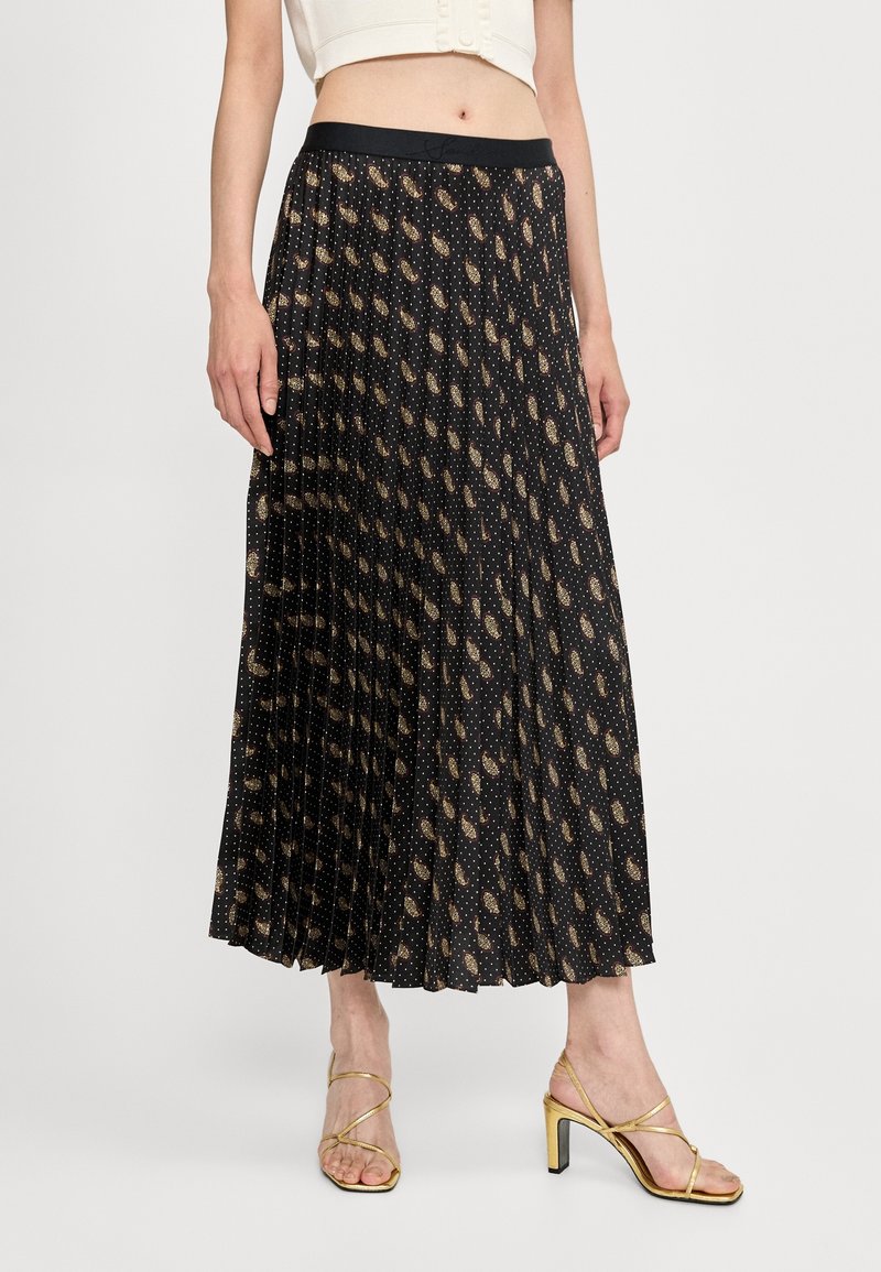 Geplooide zwarte rok met een metalen gouden paisleypatroon, met een zwarte elastische tailleband. Gecombineerd met gouden sandaaltjes met bandjes.