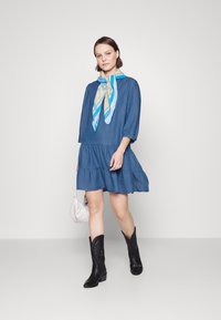 Robe en denim à manches trois-quarts, avec un ourlet à volants et une écharpe à motifs bleu. Portée avec des bottes de cowboy noires et un sac à main clair.