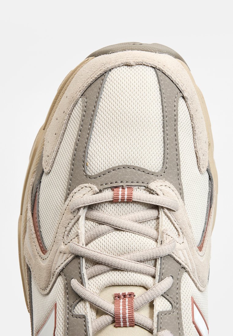 Baskets de sport beiges avec des matériaux en maille et en suède. Présente un design à lacets, des accents gris et des surfaces texturées. Logo triangulaire notable.