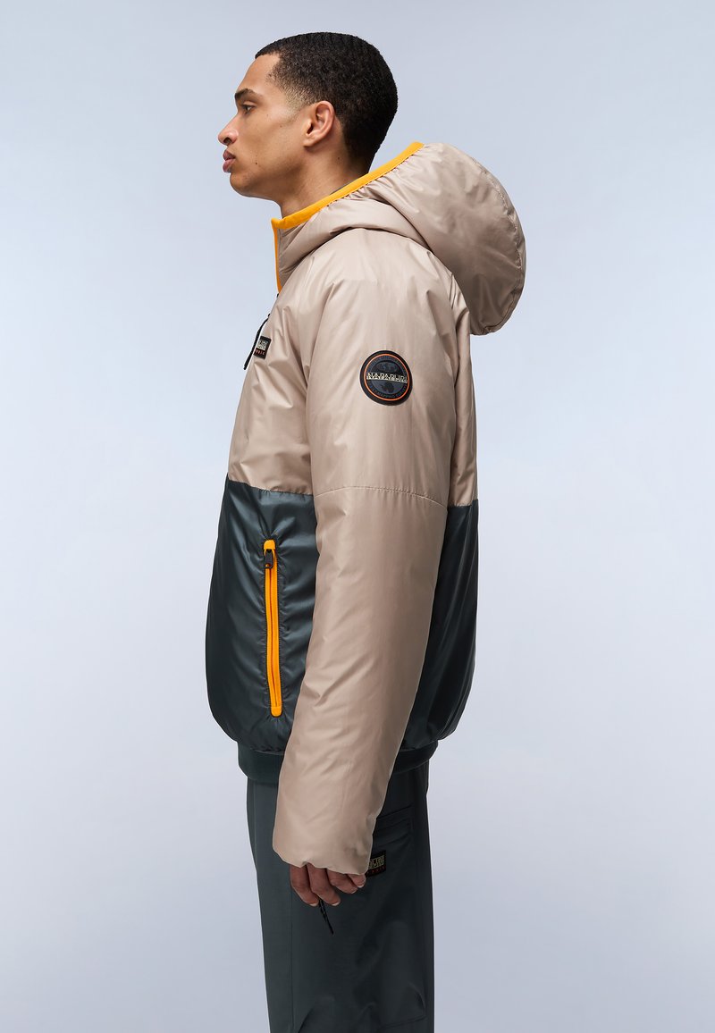 Chaqueta ligera bicolor en beige y verde oscuro, con capucha, detalles en naranja, parche con logo en la manga y bolsillo lateral con cremallera.