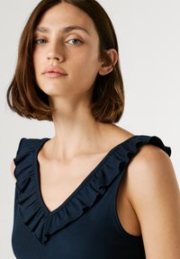 Marinblå linne med ribbad struktur, som har en djup V-ringning och volangdetaljer längs axelbanden.