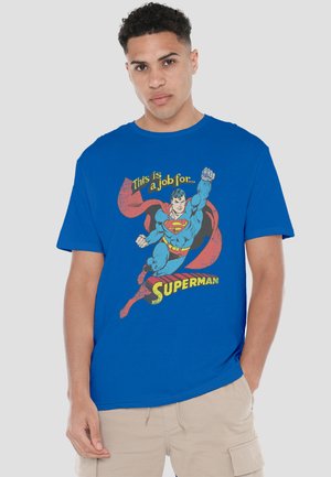 Young man with short curly hair wearing blue Superman T-shirt and beige cargo shorts, standing against plain gray background.