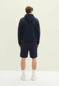 Muž stojící zády, oblečený v námořnickém mikině s kapucí, námořnických cargo šortkách, bílých ponožkách a bílých teniskách na jednotném pozadí.