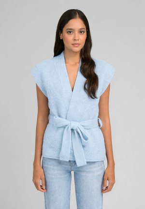 Femme aux longs cheveux foncés portant un gilet sans manches ceinturé bleu clair et un jean bleu clair, debout devant un fond gris uni.