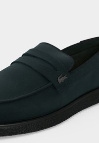 Mocassin vert foncé avec semelle noire texturée et petit logo crocodile sur la lanière latérale sur fond clair uni.