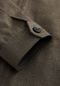 Gros plan sur la manche d'une veste en tissu brun texturé avec un bouton noir au poignet.