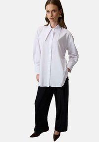 Kobieta ubrana w przew oversized biały koszulę z szerokim, spiczastym kołnierzem, czarne szerokie spodnie, buty w cętki o spiczastym nosku oraz złote kolczyki.