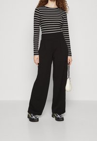 Svarta vida byxor med sidofickor, tillsammans med en svart-och-vit randig långärmad skjorta. Vardagsskor och en liten vit handväska.