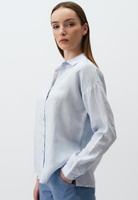 Hellblaues Leinenhemd mit durchgehender Knopfleiste, klassischem Kragen, langen Ärmeln und subtiler Textur. Getragen mit hellblauen Jeans.