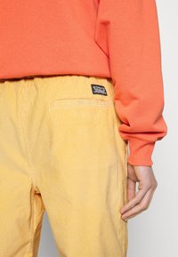 Pomarańczowy sweatshirt z prążkowanymi mankietami, w połączeniu z żółtymi spodniami z sztruksu, które mają widoczną tylną kieszeń i metkę z logo marki.