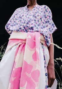 Person klädd i en blommig blus instoppad i vita byxor med en rosa och beige stickad tröja knuten runt midjan mot en mörk bakgrund.