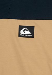 Šviesiai rudos spalvos audinys su tekstūriniu paviršiumi, turintis tamsiai mėlyną plokštelę ir juodą Quiksilver logotipo uždėtą detalę su baltais akcentais.