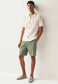 Chemise texturée beige clair à manches courtes avec des boutons marron, associée à un short vert olive et des chaussures slip-on beige clair. Style simple et décontracté.