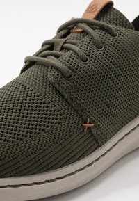 Sneaker intrecciati verdi con superficie testurizzata, cuciture a contrasto in marrone e suola in gomma grigia. I lacci e i fori per i lacci completano il design.