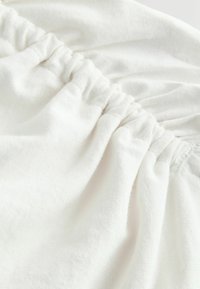 Weißer Baumwollstoff mit geraffter Textur, der eine weiche, leicht gerippte Oberfläche und elastische Raffung am Rand zeigt. Keine Muster oder Akzente sichtbar.