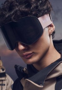 Smučarska očala z veliko, ukrivljeno črno lečo, rožnatim trakom z napisom "HAY" in mehko teksturo, ki jih nosi oseba v temni jakni.