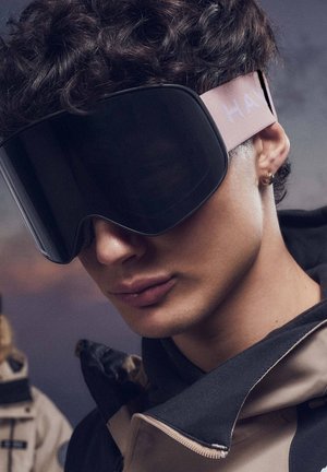 Ski goggles with a large, curved black lens, pink strap featuring "HAY" text, and a soft texture, worn by a person in a dark jacket.