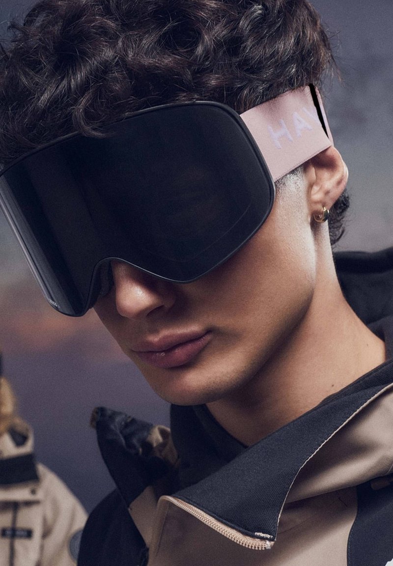 Smučarska očala z veliko, ukrivljeno črno lečo, rožnatim trakom z napisom "HAY" in mehko teksturo, ki jih nosi oseba v temni jakni.