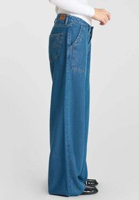 Jeans de denim de gran tamaño color azul claro con una cintura alta, grandes bolsillos y un parche con logo visible en la parte trasera. La tela tiene una textura suave.