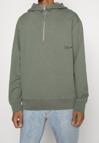 Sudadera con cremallera en verde oliva, con un bolsillo tipo canguro y puños de canalé. Presenta el logotipo "REPLAY" en negro en la parte izquierda del pecho. Tejido suave.
